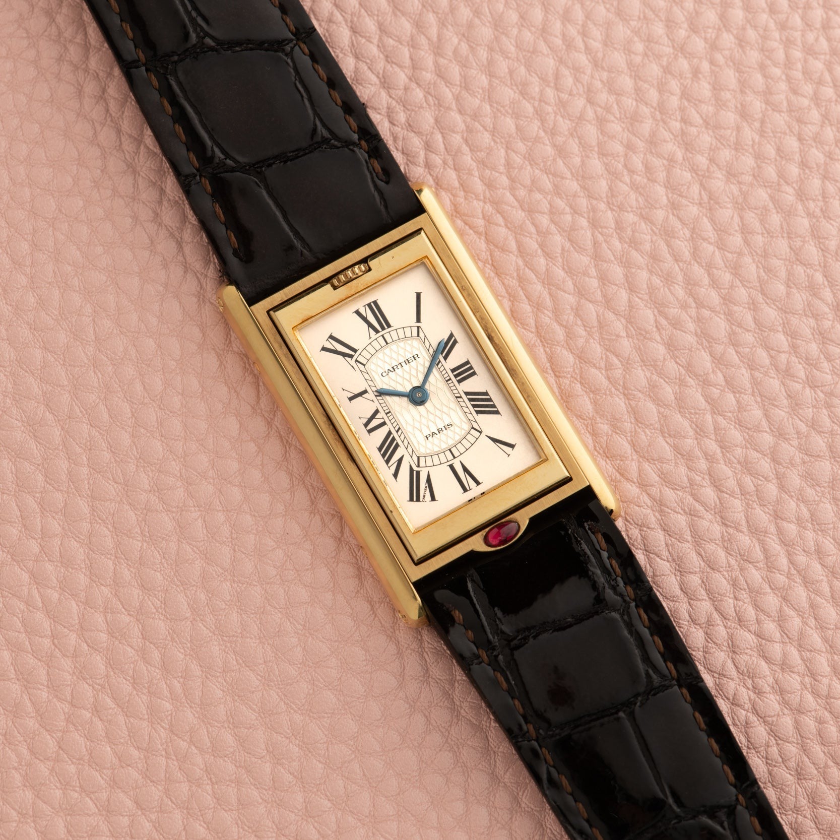 The Cartier Tank Basculante Mécanique 150th Anniversary Limited Edition, a rectangular gold watch with Roman numerals and a black leather strap, displayed on a pink textured surface.