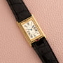 The Cartier Tank Basculante Mécanique 150th Anniversary Limited Edition, a rectangular gold watch with Roman numerals and a black leather strap, displayed on a pink textured surface.
