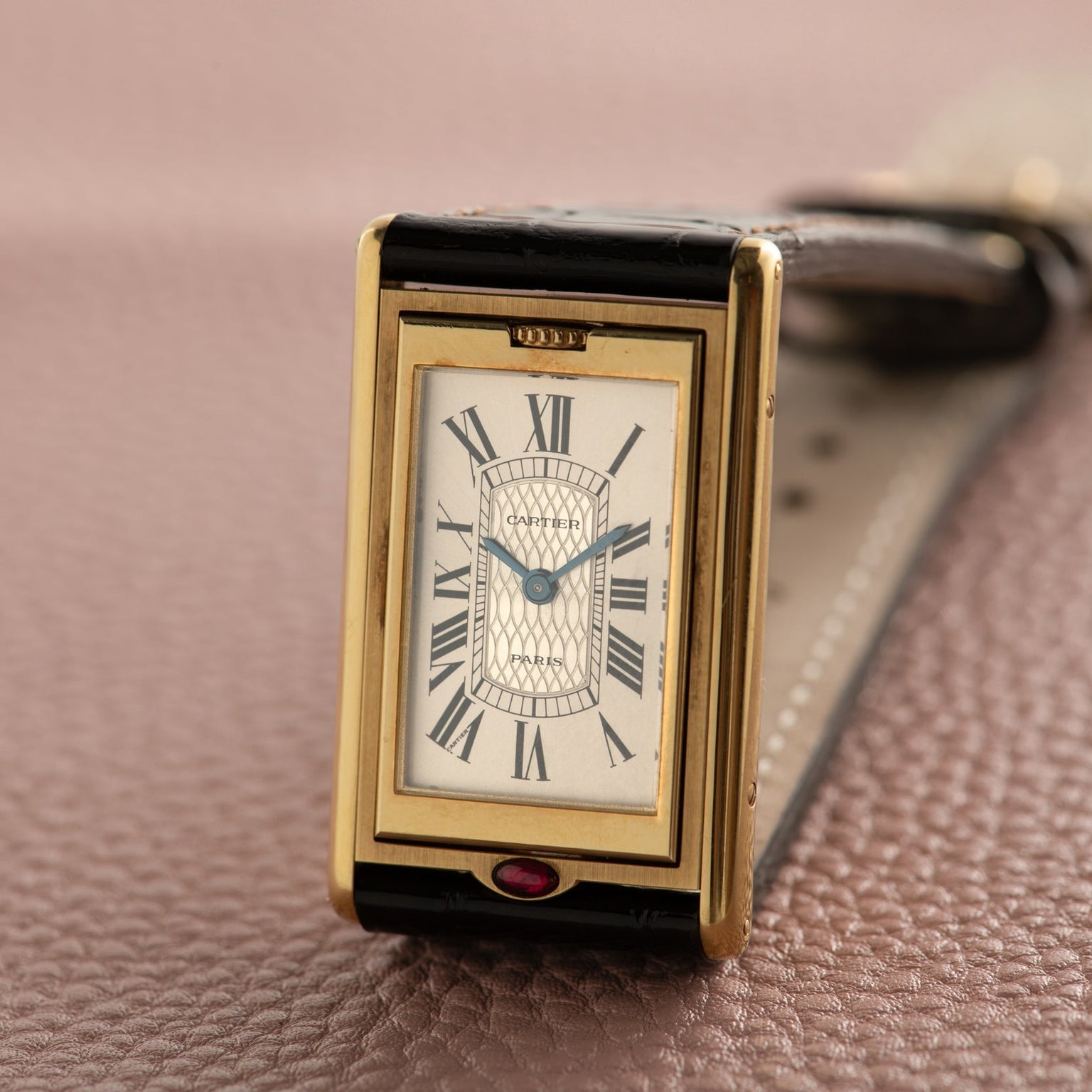 The Cartier Tank Basculante Mécanique 150th Anniversary Limited Edition is a gold rectangular wristwatch with Roman numerals, displayed on a textured brown surface.