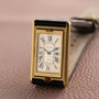 The Cartier Tank Basculante Mécanique 150th Anniversary Limited Edition is a gold rectangular wristwatch with Roman numerals, displayed on a textured brown surface.