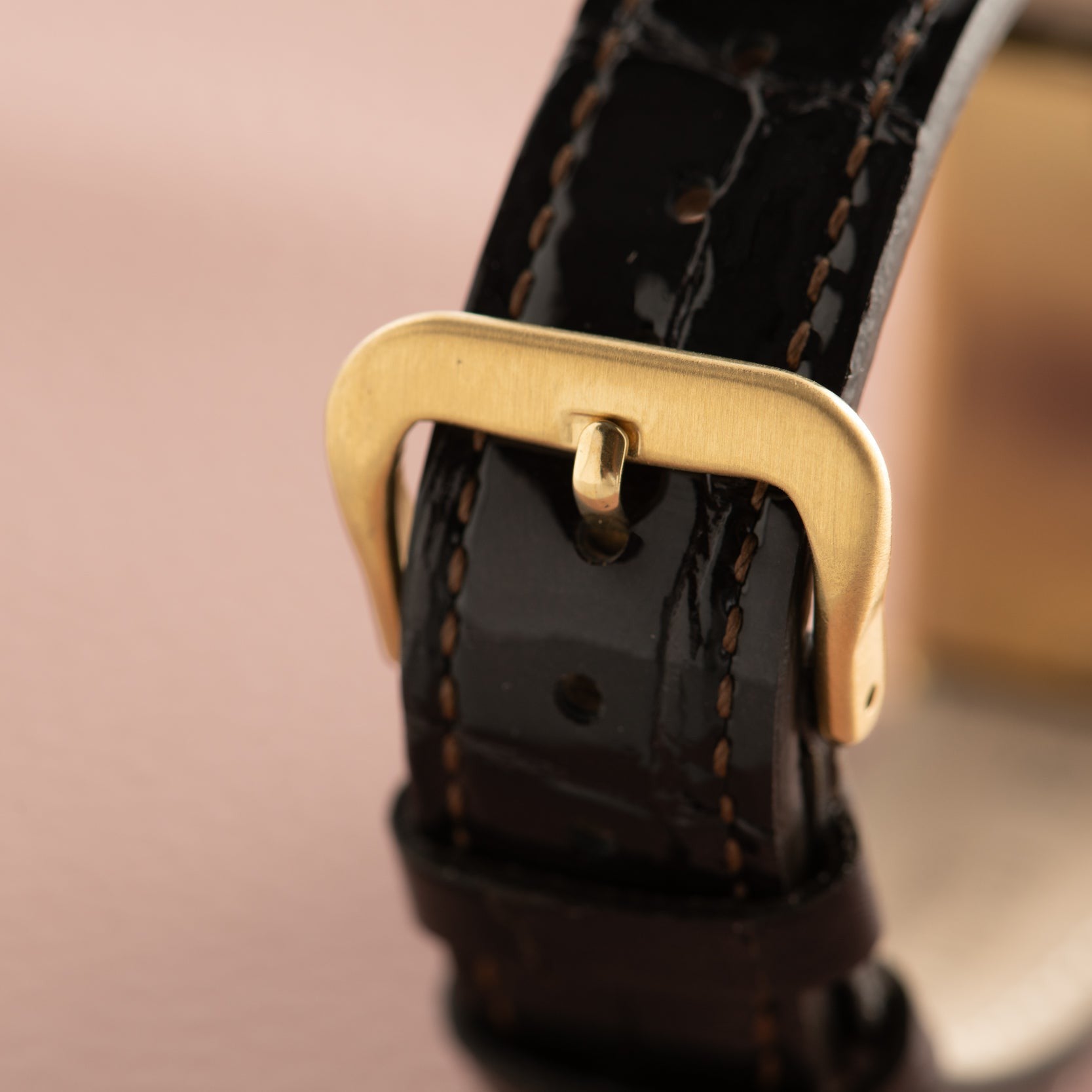 Close-up of the Cartier Tank Basculante Mécanique 150th Anniversary Limited Editions black leather strap with a gold buckle, displayed on a soft pink background.