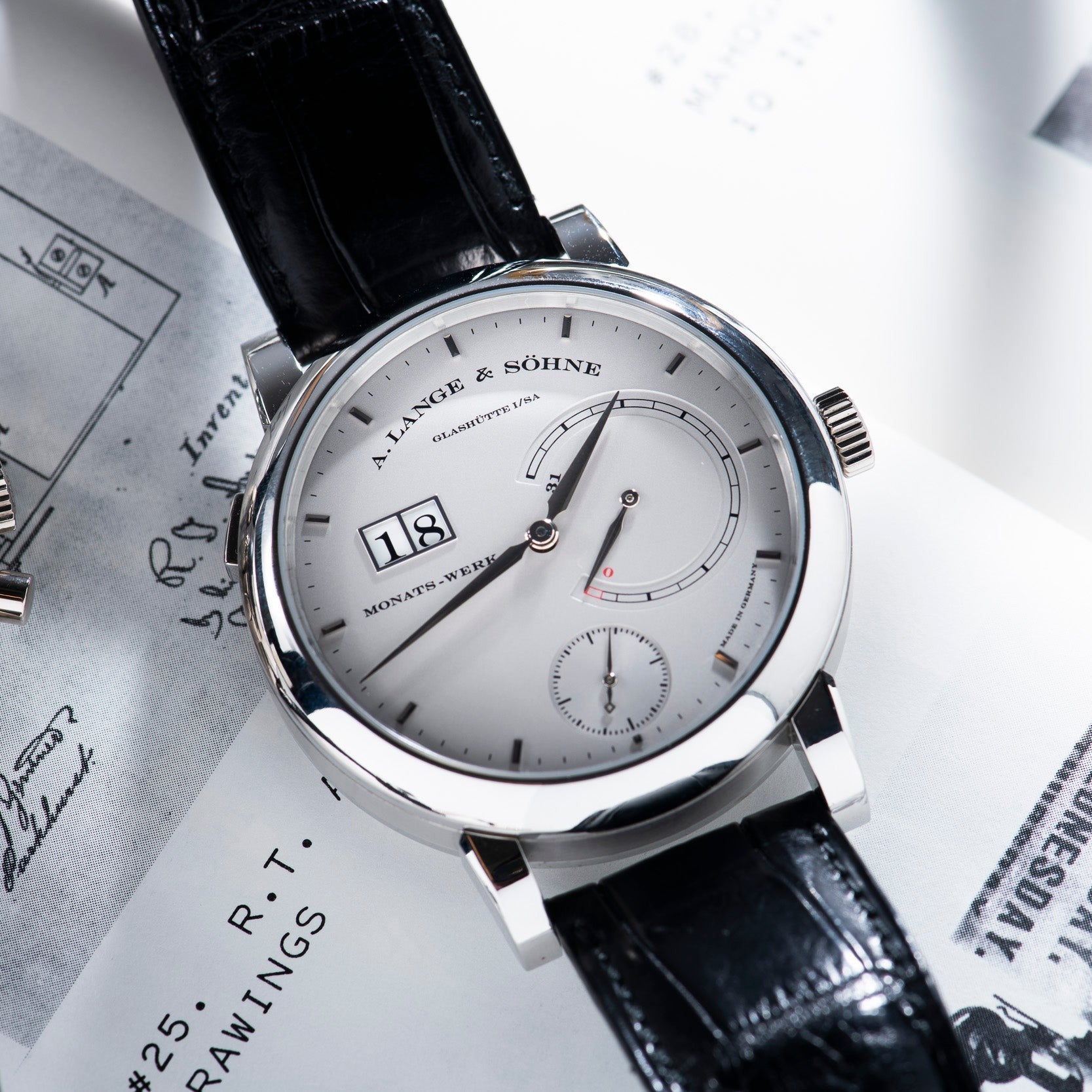 A Lange & Sohne Lange 31 wristwatch featuring a silver dial and black leather strap, displayed on a white paper background.