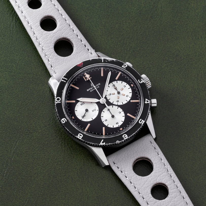 A Breitling Co-Pilot Chronograph wristwatch in black and silver features a perforated gray leather strap, displayed against a green background.