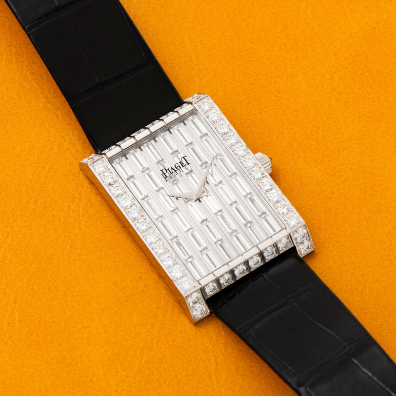 The Piaget Limelight Baguette Diamond Dress Watch, featuring a rectangular case and black strap, is displayed on an orange surface.