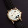 The Lange & Söhne Lange 1 Tourbillon features a gold case, leather strap, and visible tourbillon, elegantly displayed on a dark surface.