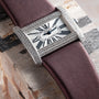 A Cartier Tank Divan watch with a diamond bezel, Roman numerals, and a purple strap rests on a wooden surface.