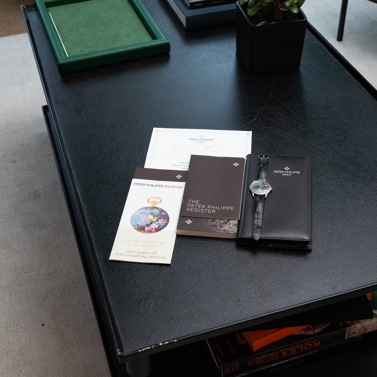 A Patek Philippe Calatrava wristwatch and branded booklets are neatly arranged on a black table.