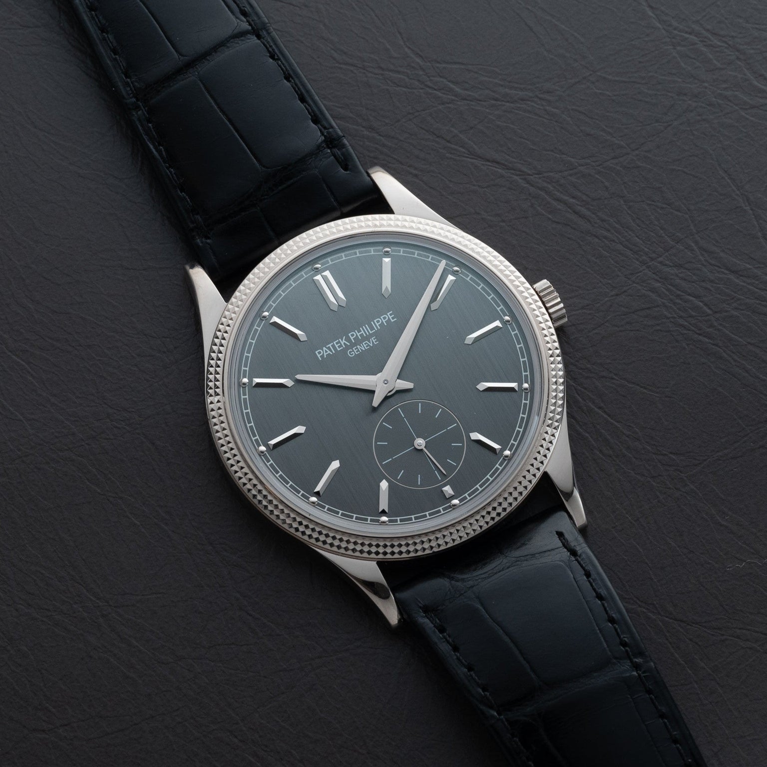 A Patek Philippe Calatrava wristwatch with a silver case and black leather strap displayed on a dark leather surface.