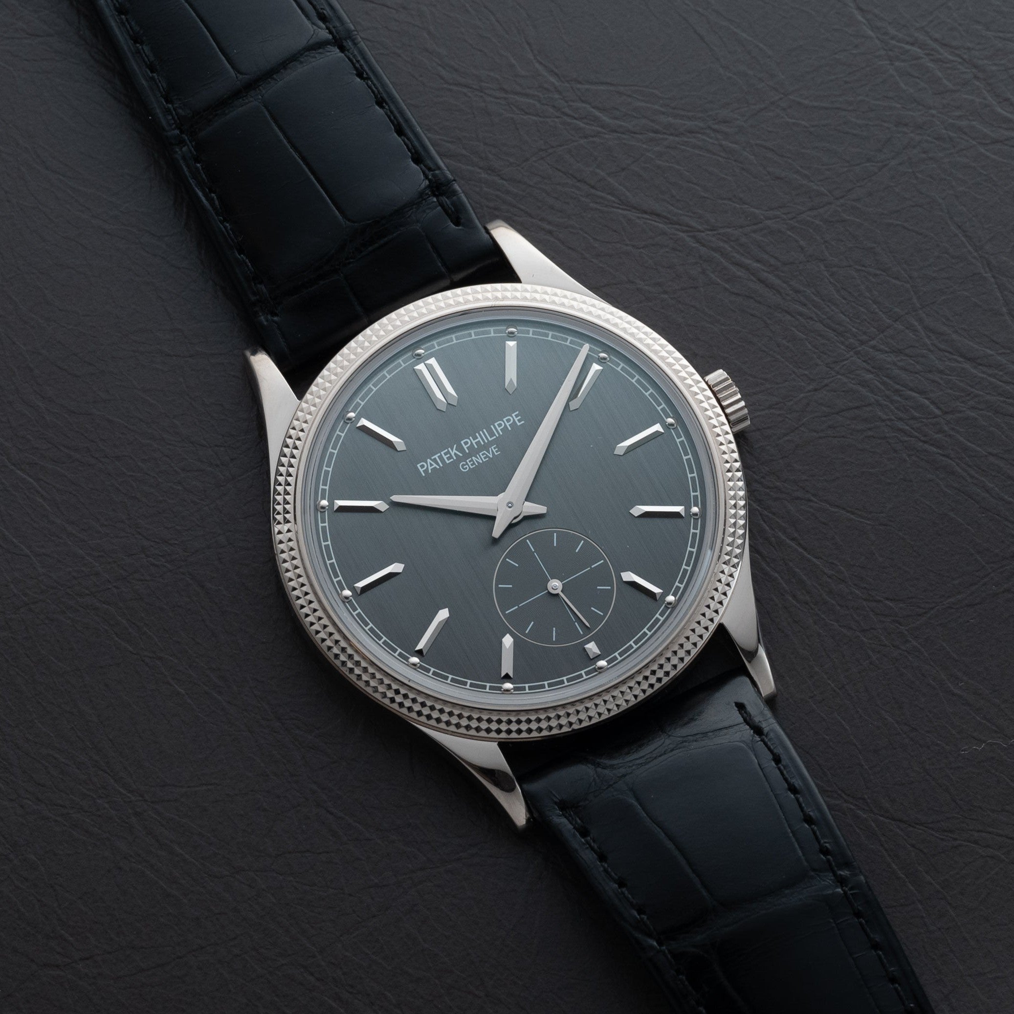 A Patek Philippe Calatrava wristwatch with a silver case and black leather strap displayed on a dark leather surface.