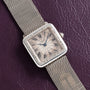 A Cartier Santos Platinum square silver watch with a mesh band and Roman numerals, displayed on a textured dark purple surface.