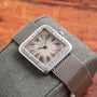 The Cartier Santos Platinum, a vintage wristwatch with Roman numerals, is displayed on a textured gray cushion atop a wooden surface.