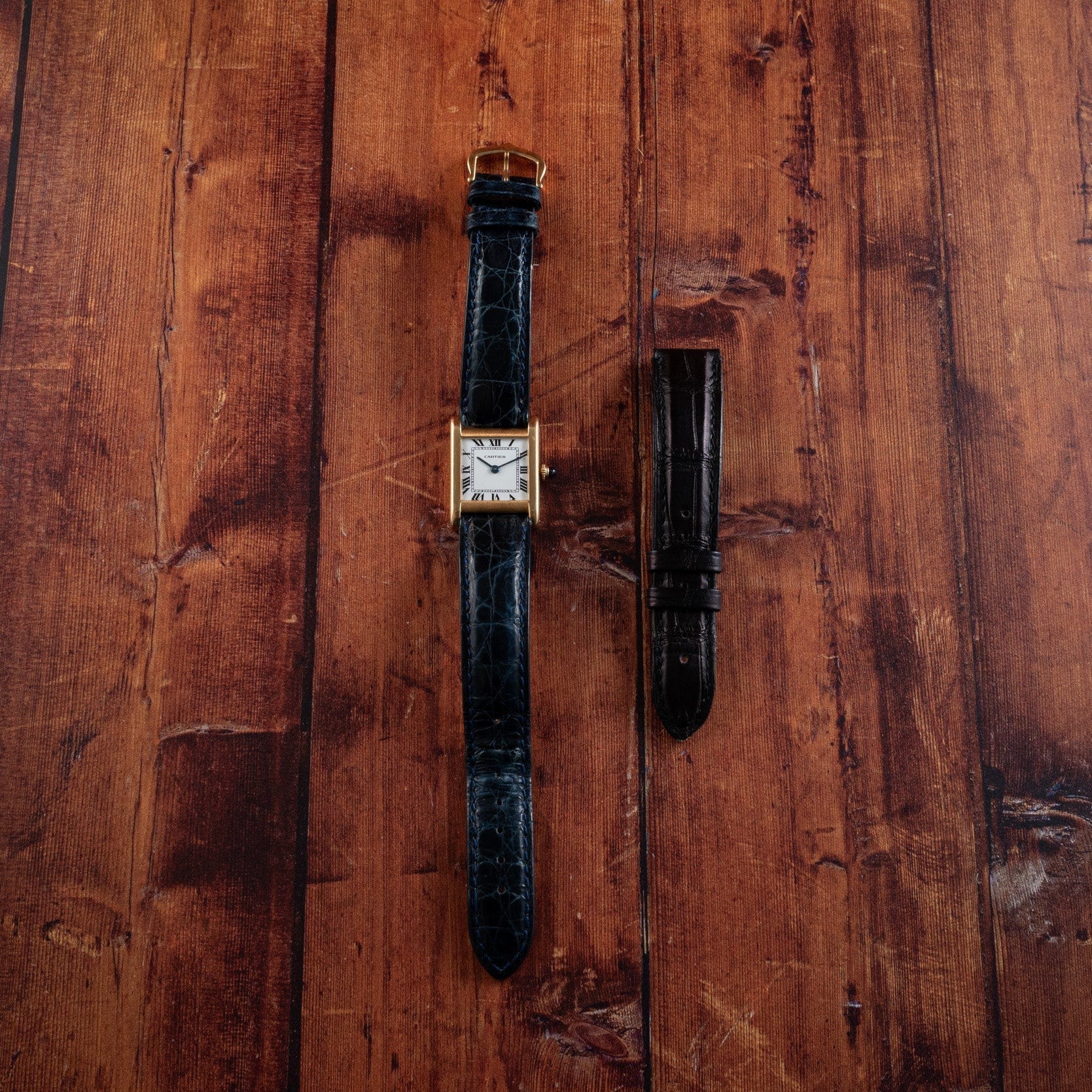 A Cartier Tank Normale wristwatch with a black strap is placed beside an additional black strap on a wooden surface.