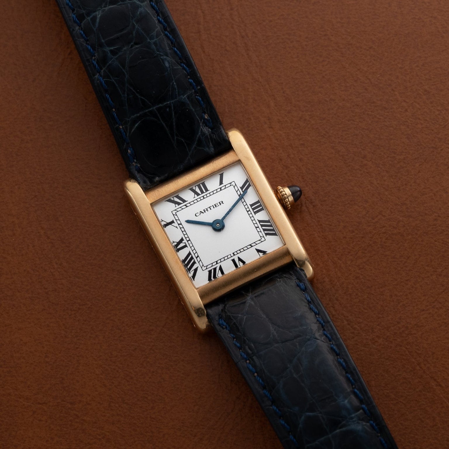 The Cartier Tank Normale watch features Roman numerals on a white dial and a black leather strap, displayed on brown leather.