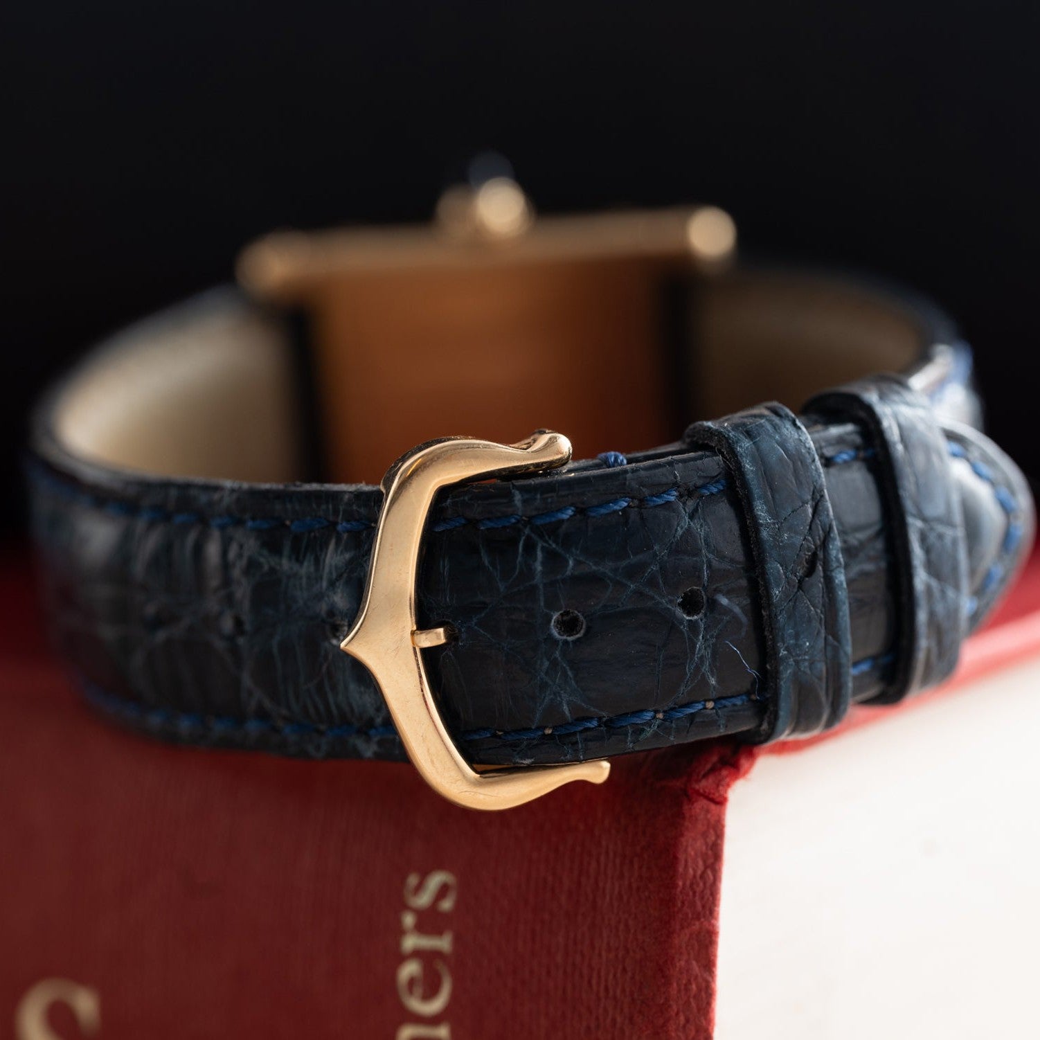 Close-up of a Cartier Tank Normale blue leather watch strap with gold buckle, resting on a red hardcover book.