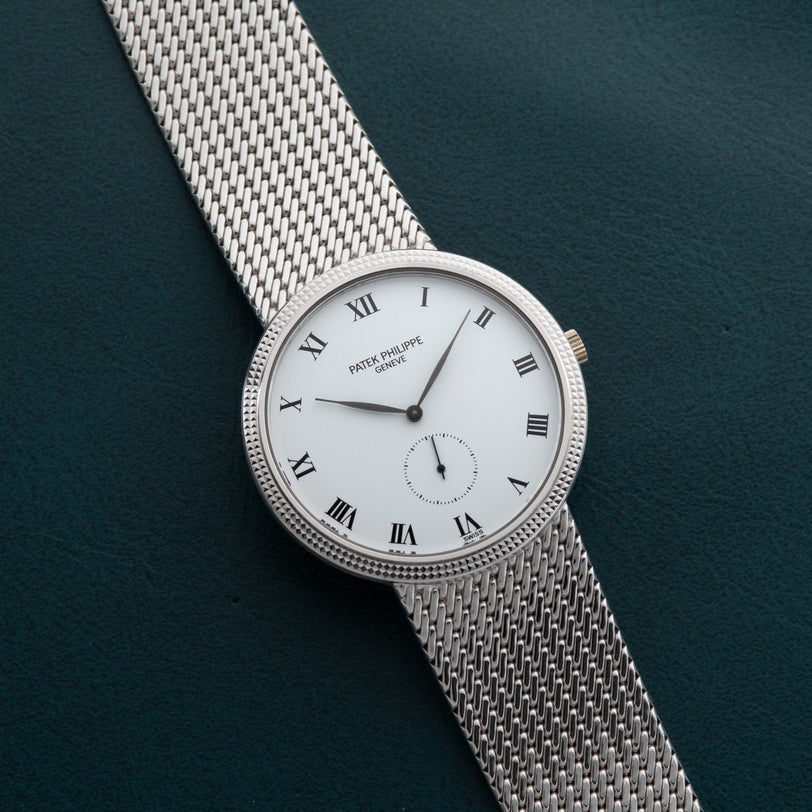 Patek Philippe Calatrava silver mesh watch with white face, Roman numerals, and classic design displayed on a dark green textured surface.