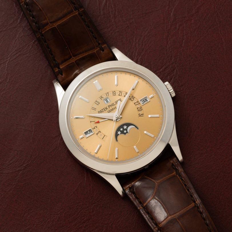 The Patek Philippe Retrograde Perpetual Calendar is a luxury wristwatch featuring a gold dial, brown leather strap, and displays for the calendar and moon phase, shown on a brown surface.
