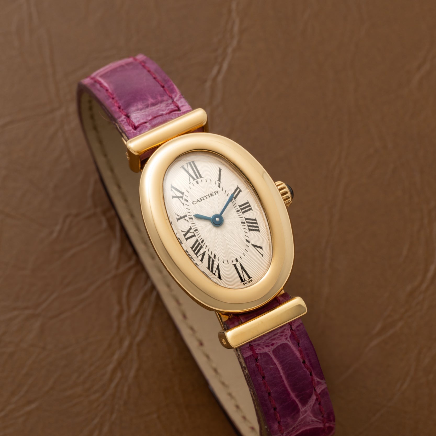 Cartier Baignoire watch with gold oval case, roman numerals, and a purple leather strap placed on a brown textured surface.