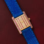The Cartier Tank Louis Diamonds watch features a jeweled face, gold case, blue strap, and pink accents, elegantly displayed on burgundy leather.
