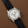 The Patek Philippe Annual Calendar Moonphase, in white gold with a white dial and black leather strap, features a moon phase display plus annual calendar with day, date, and month indicators—all set on a brown leather surface.