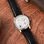 The Lange & Sohne Lange 1 Tourbillon Perpetual Calendar, featuring a black leather strap and silver dial, is elegantly displayed on a wooden surface.