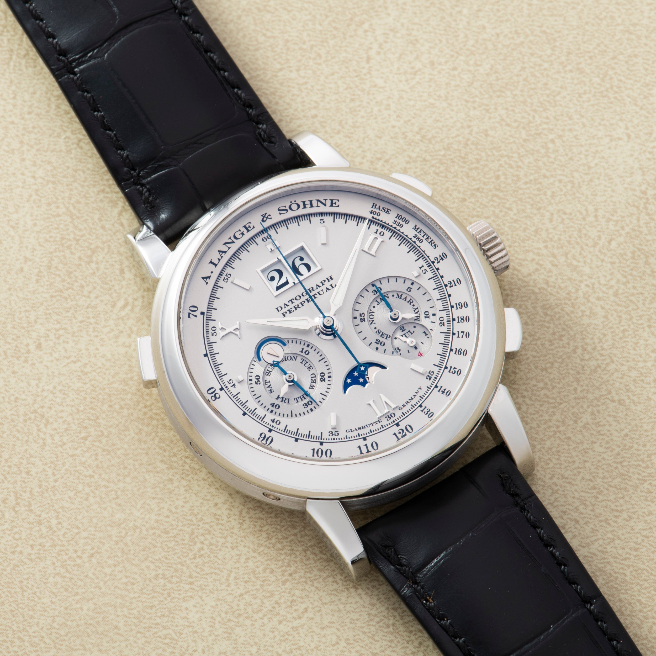A luxurious Lange & Sohne Datograph Perpetual Calendar wristwatch with a black leather strap and multiple dials, displayed on a beige surface.