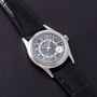 A Patek Philippe Calatrava wristwatch in silver with a black leather strap, displayed on a dark leather surface.