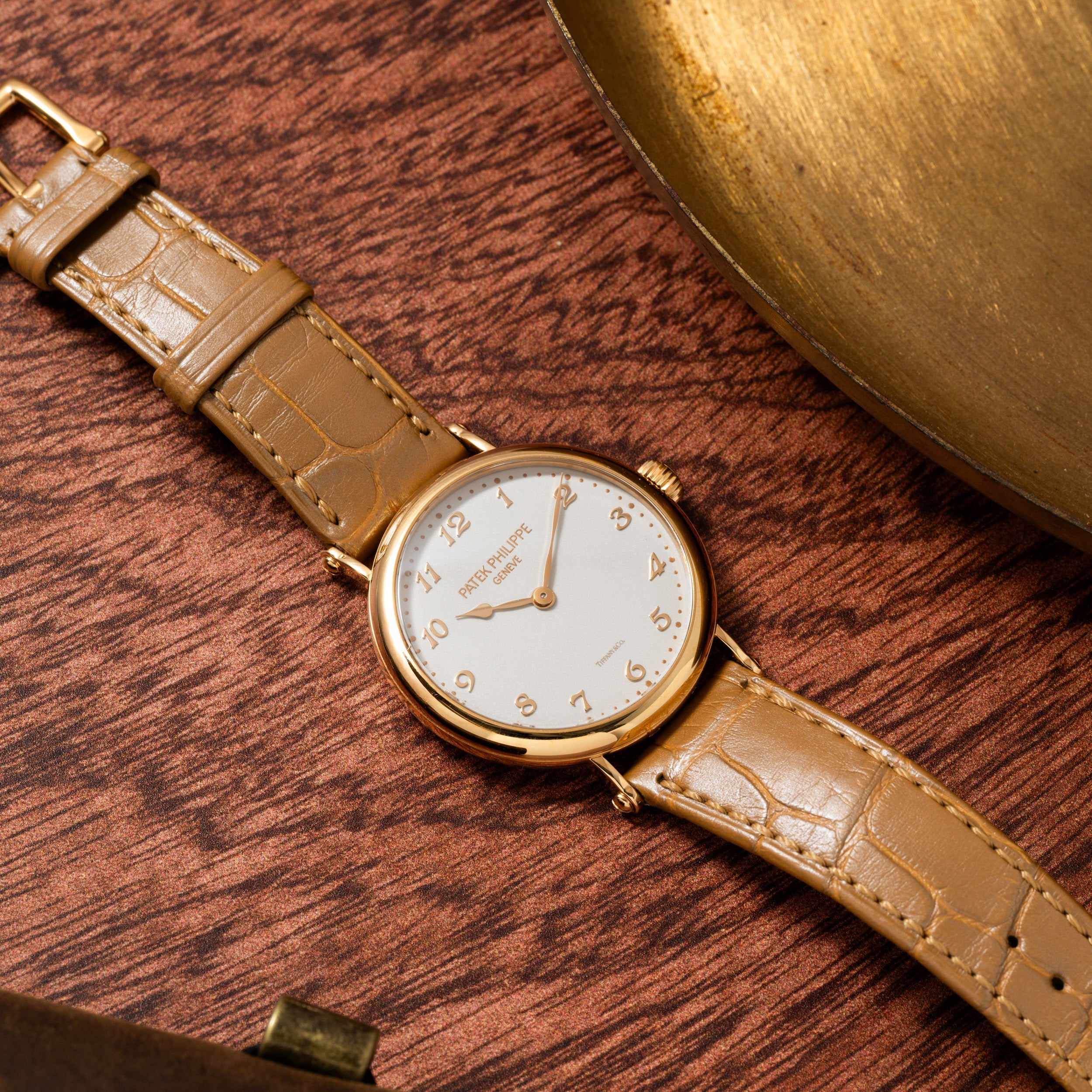 The Patek Philippe Calatrava, featuring a gold case and tan leather strap, rests on a wooden surface beside a brass object.