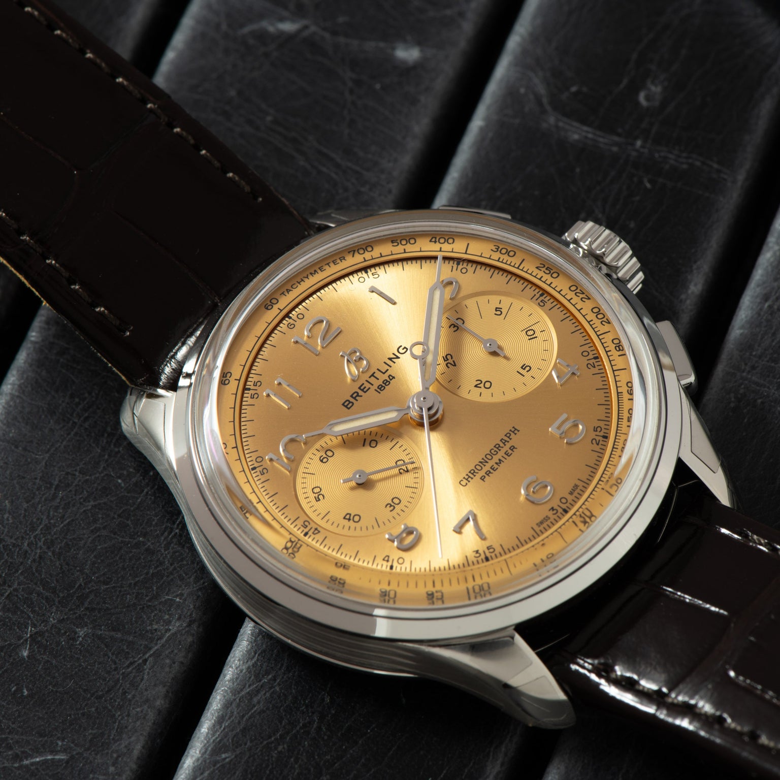 The Breitling Premiere B09 Chronograph 40 features a gold dial and brown leather strap, displayed on a black textured surface.