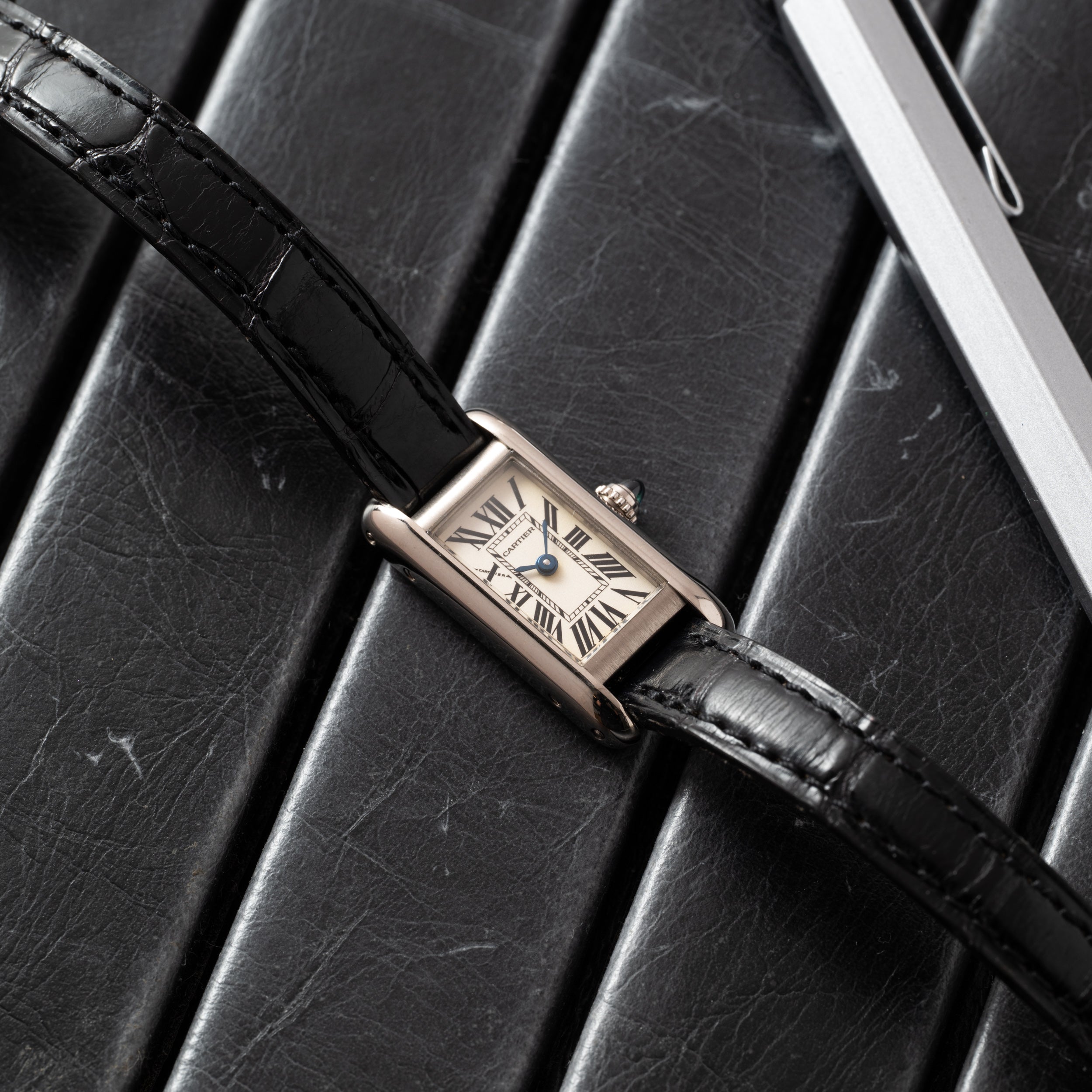 A Cartier Tank Ladies Allongee wristwatch with a black leather strap rests on a black leather surface near a silver pen.