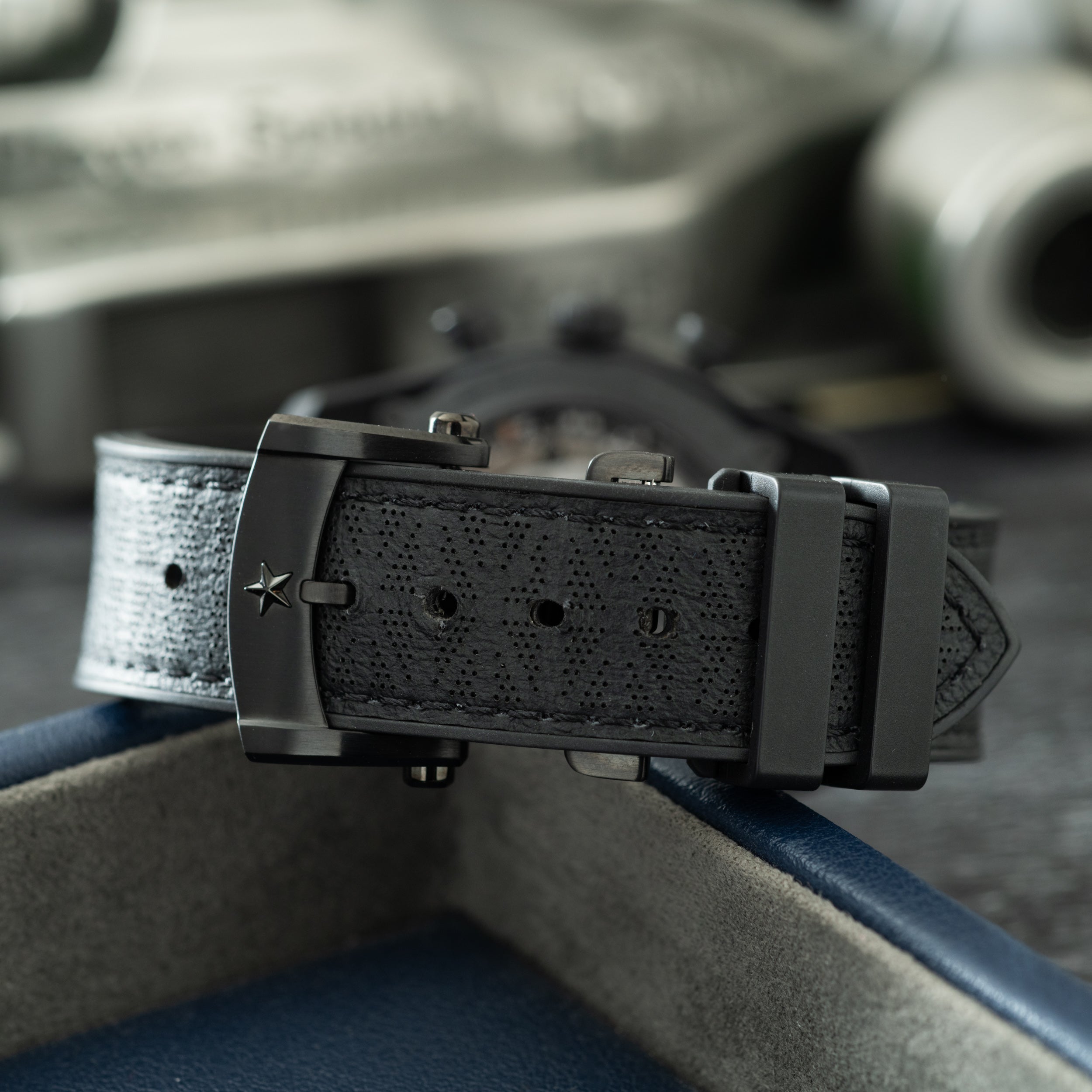 Black perforated rubber watch strap with gunmetal buckle and star emblem, displayed on blue fabric.