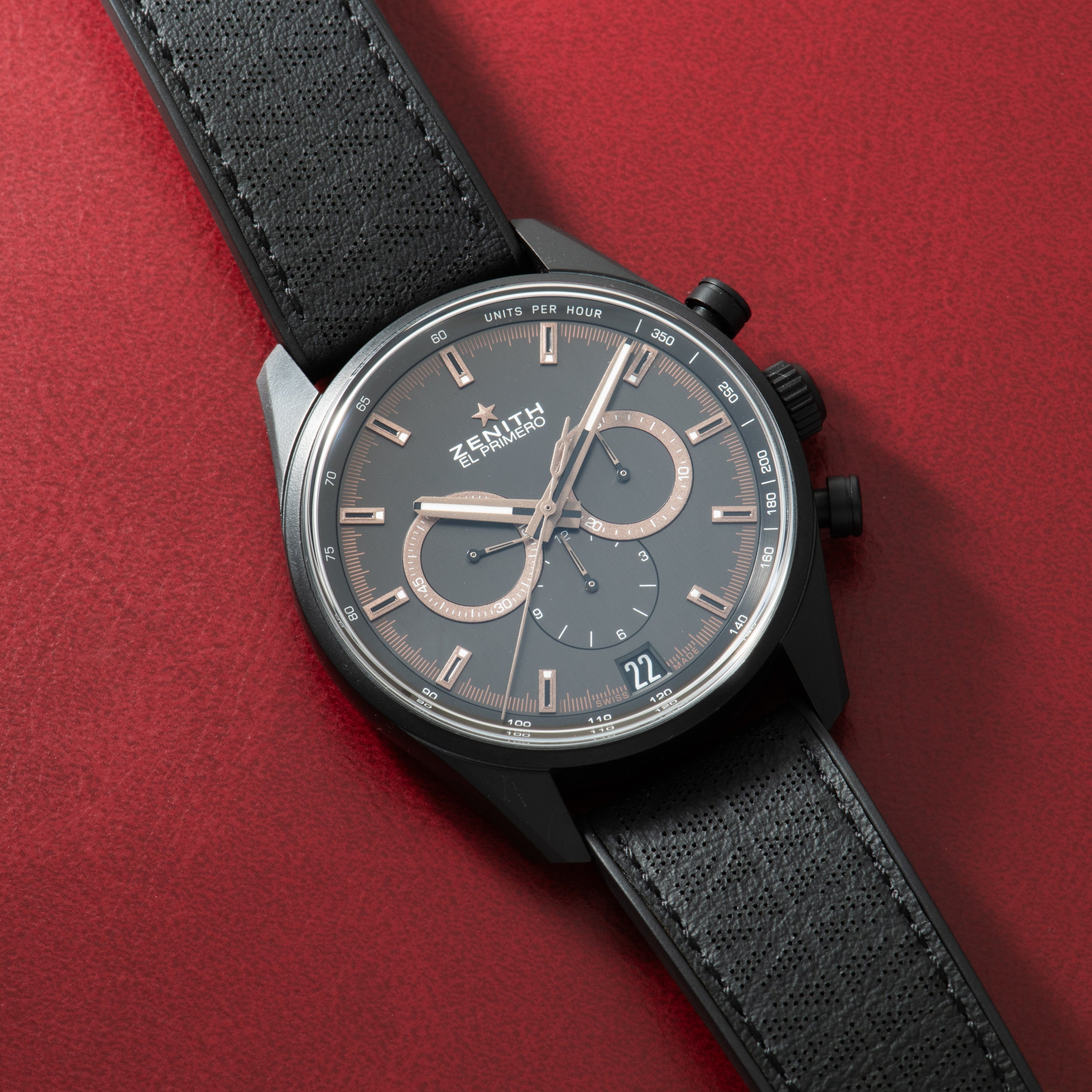 Zenith chronograph watch with dark gray dial, rose gold indices, two subdials, and black textile strap on red background.