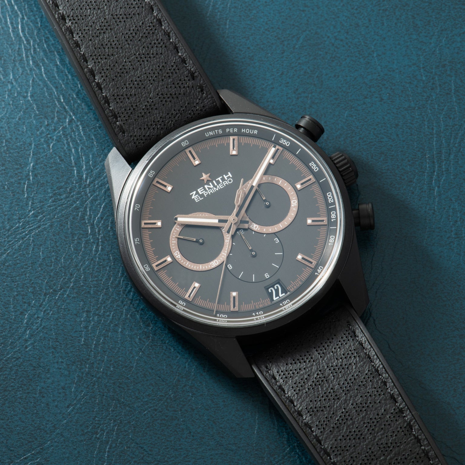 Zenith chronograph watch with gunmetal dial, rose gold accents, dual subdials, and black textile strap on teal background.