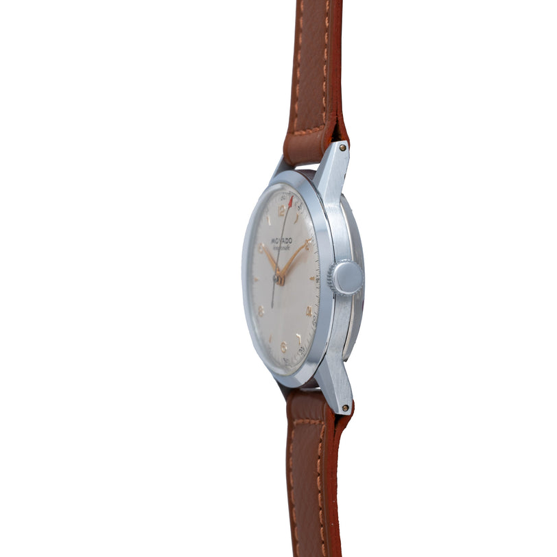 Stainless steel watch with cream dial, gilt hands, Arabic numerals, and brown leather strap with stitching.