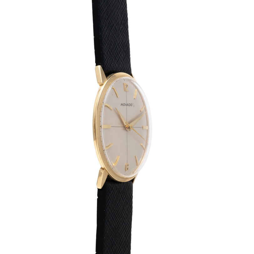 Gold-tone Movado watch with silver dial, gold hour markers, and black fabric strap.