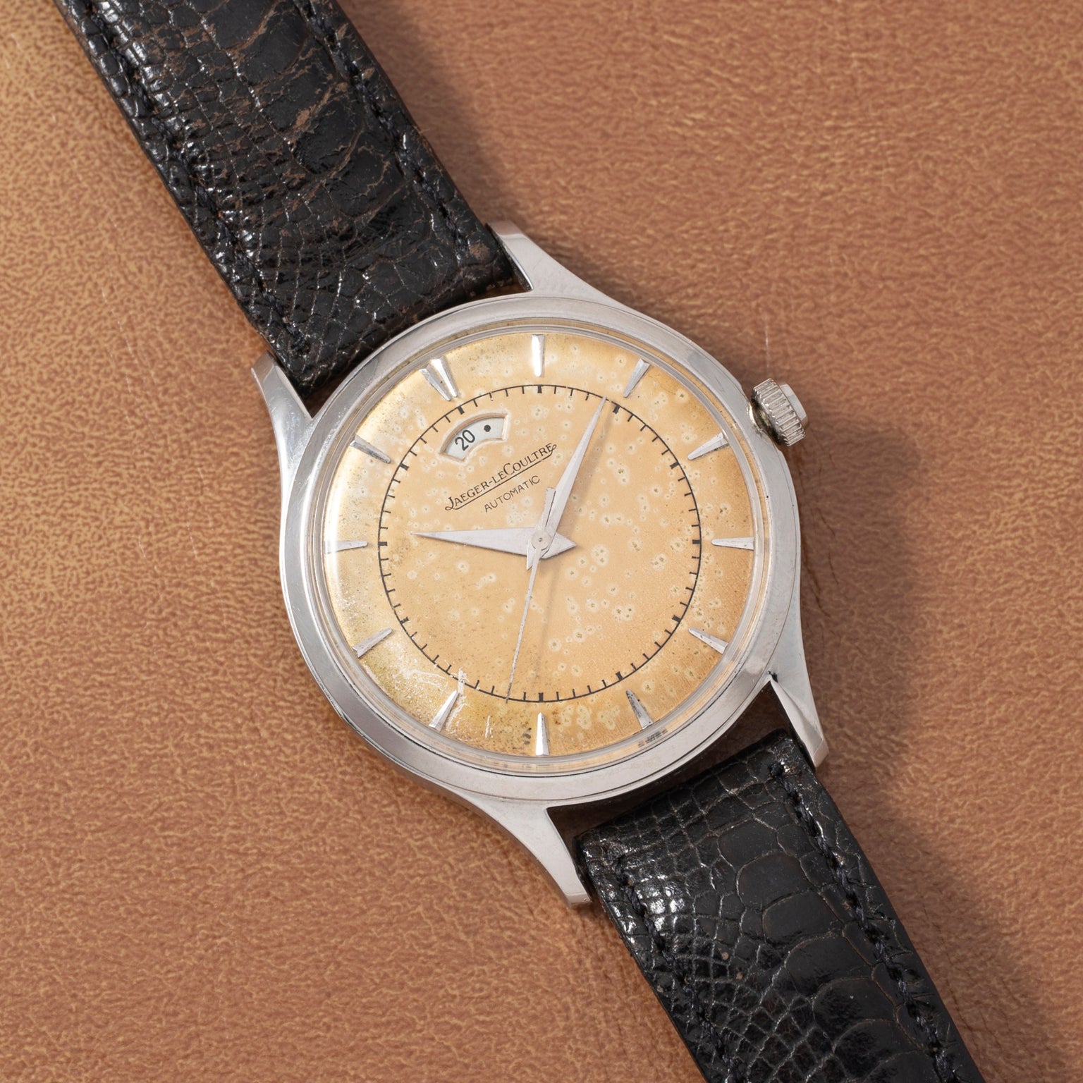 A Jaeger LeCoultre 'European Market' Powermatic wristwatch with a patinated beige dial, silver indices, date window, power-reserve display, and black leather strap is showcased on a brown textured surface.