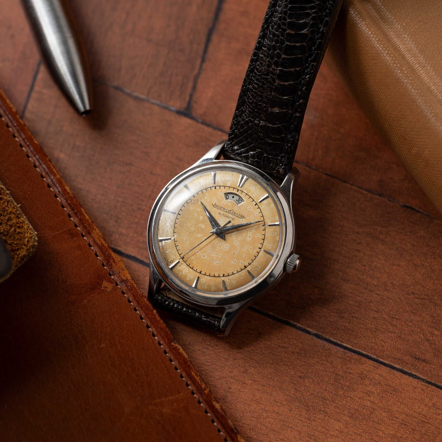 A vintage Jaeger LeCoultre 'European Market' Powermatic with a beige dial and black leather strap rests on wood beside a silver pen and brown leather item—a timeless tribute to classic midcentury watch design.
