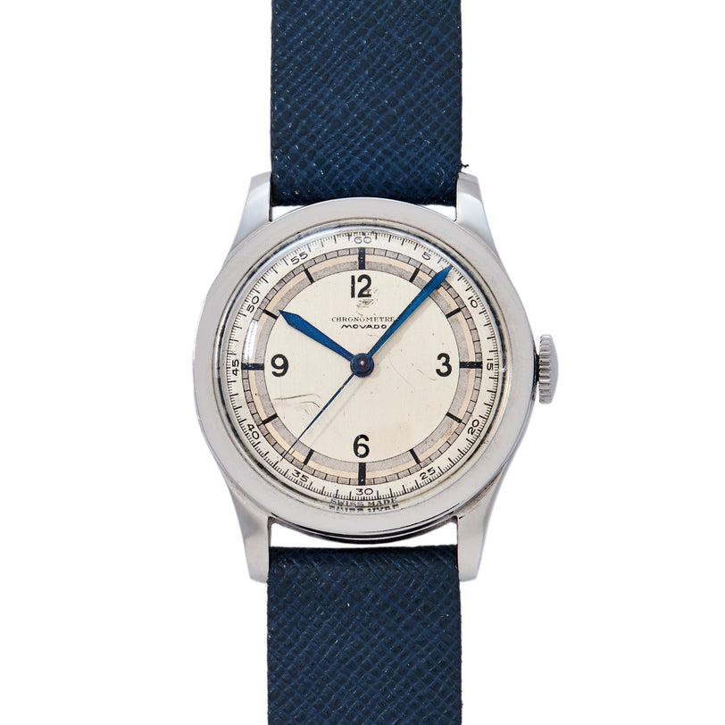 The Movado 'Calatrava' Steel is a vintage mid-century mechanical watch with a round silver case, off-white dial, black Arabic numerals, blue hands, central seconds hand, outer minute track, and blue textured leather strap.