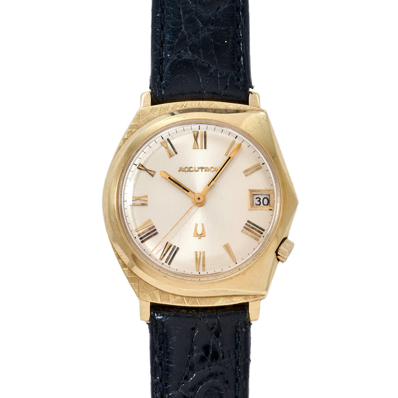 Gold-tone Bulova Accutron watch with cream dial, Roman numerals, date window, and black leather strap.