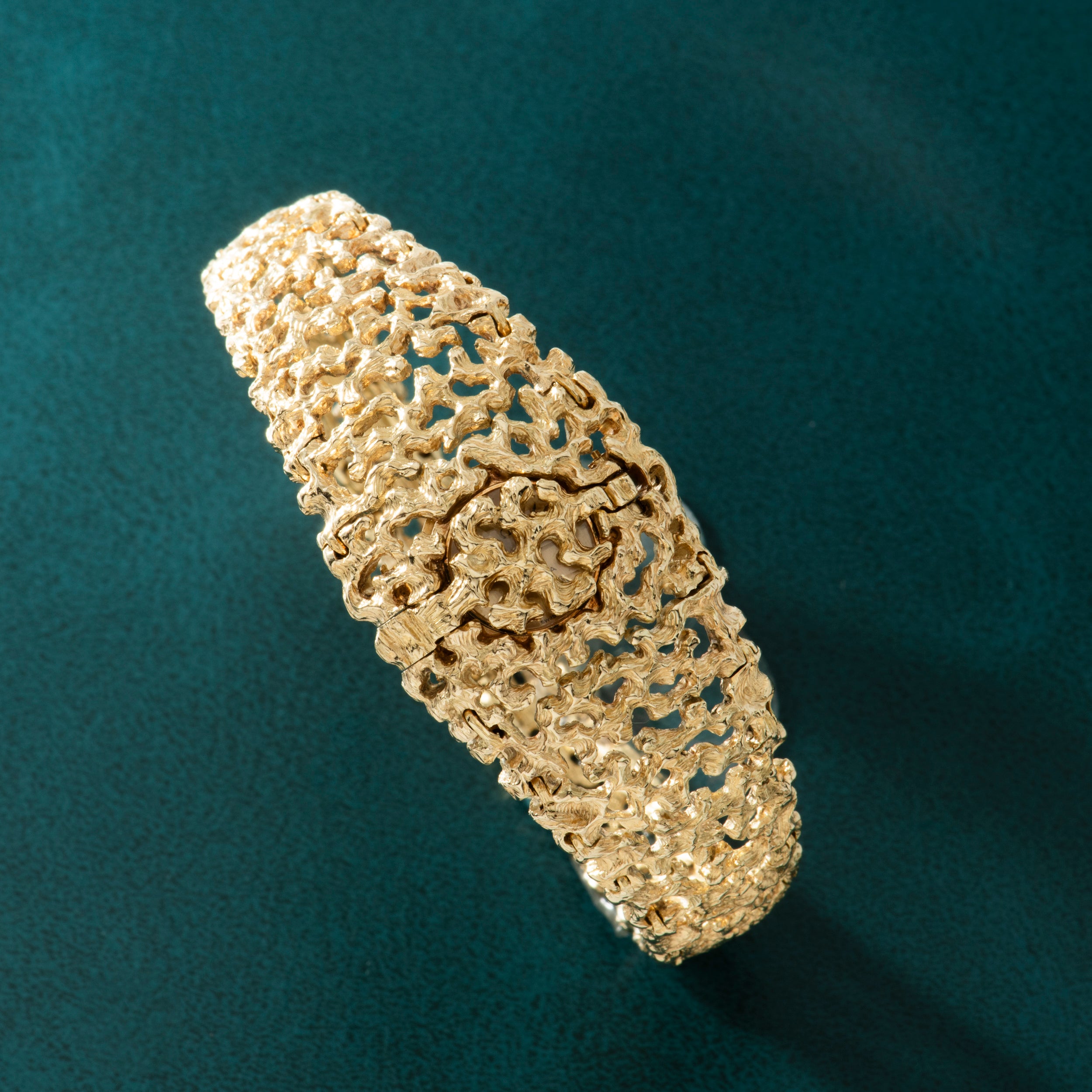 Gold-tone bracelet watch with textured organic surface pattern and concealed dial, displayed at an angle on teal background.