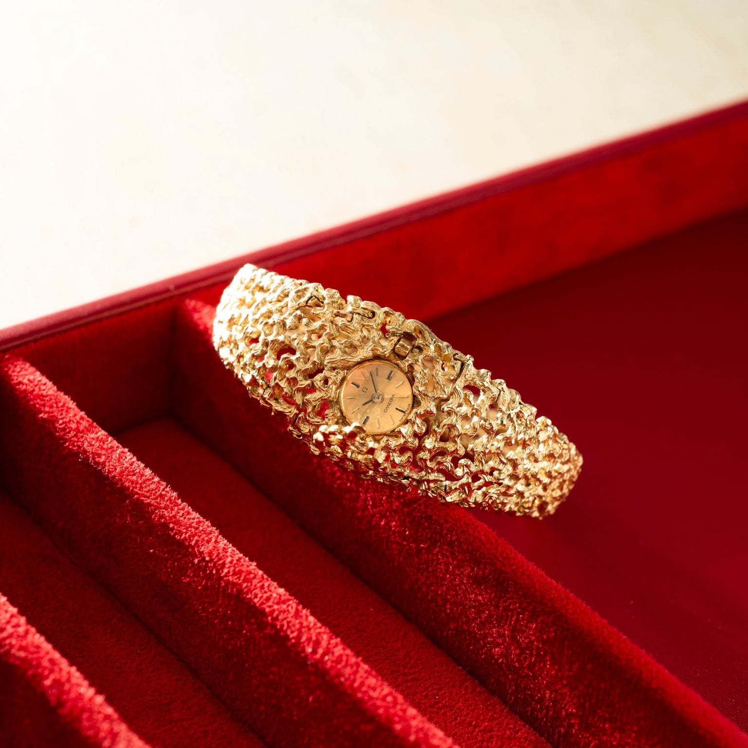 Gold-tone textured bracelet watch with small round dial, displayed in red velvet case.