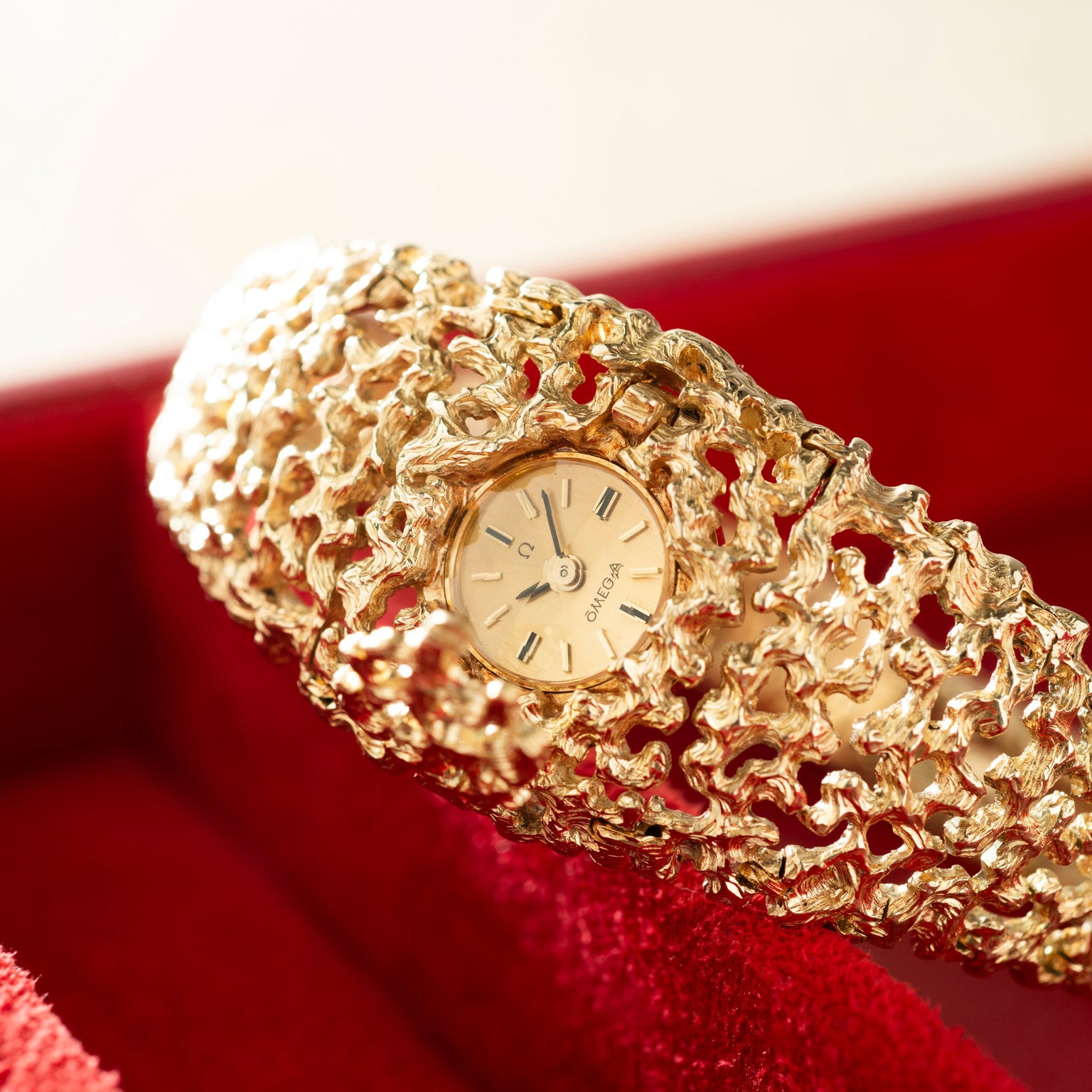 Gold textured bracelet watch with round cream dial and red gemstone accents on red fabric background.