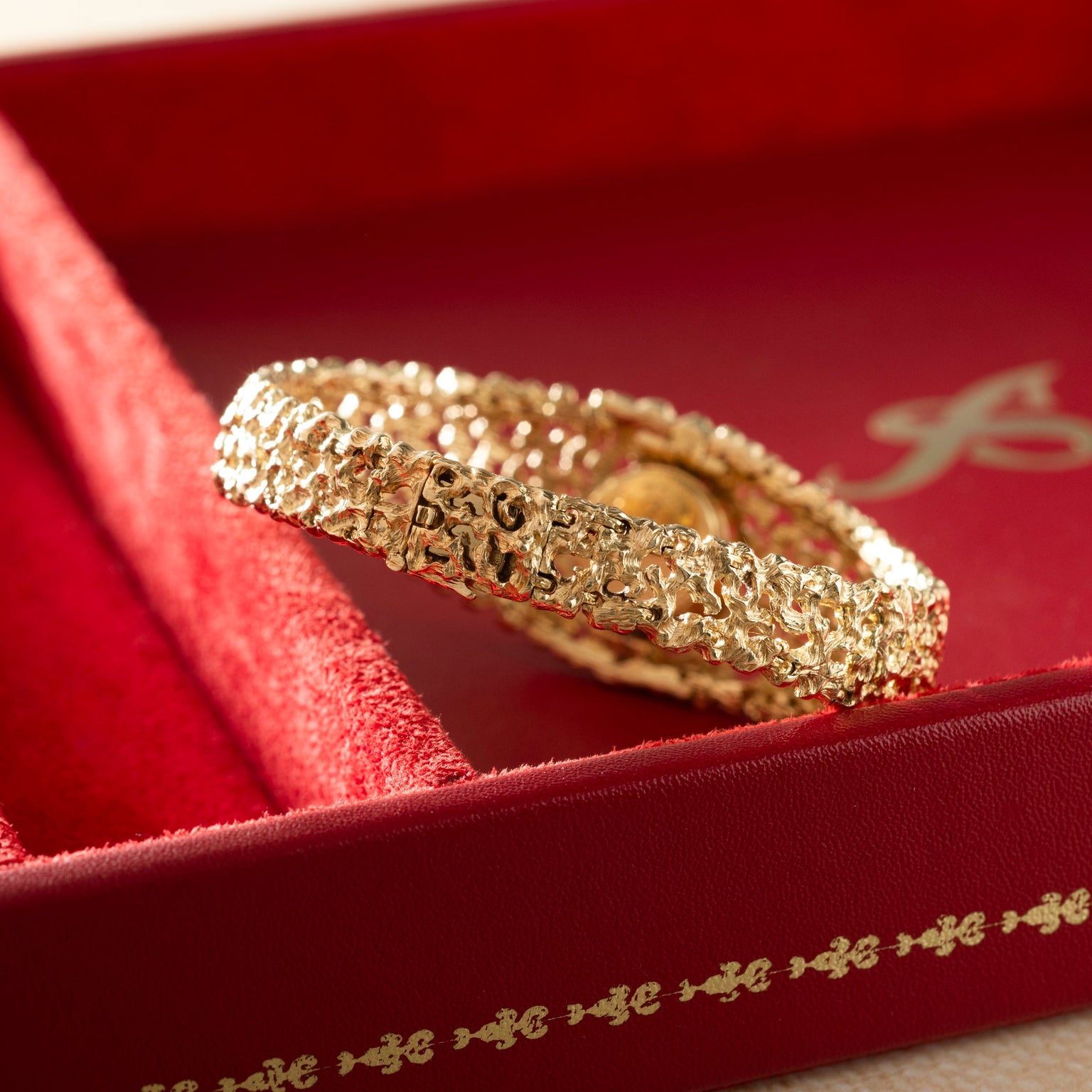 Gold-toned textured bracelet watch with integrated dial displayed on red fabric presentation box.