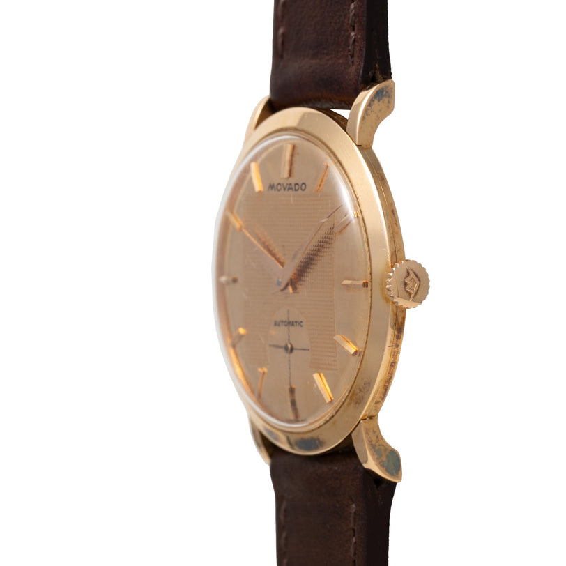 Movado Gentleman wristwatch with gold-tone oval case, champagne dial, applied indices, and dark brown leather strap.