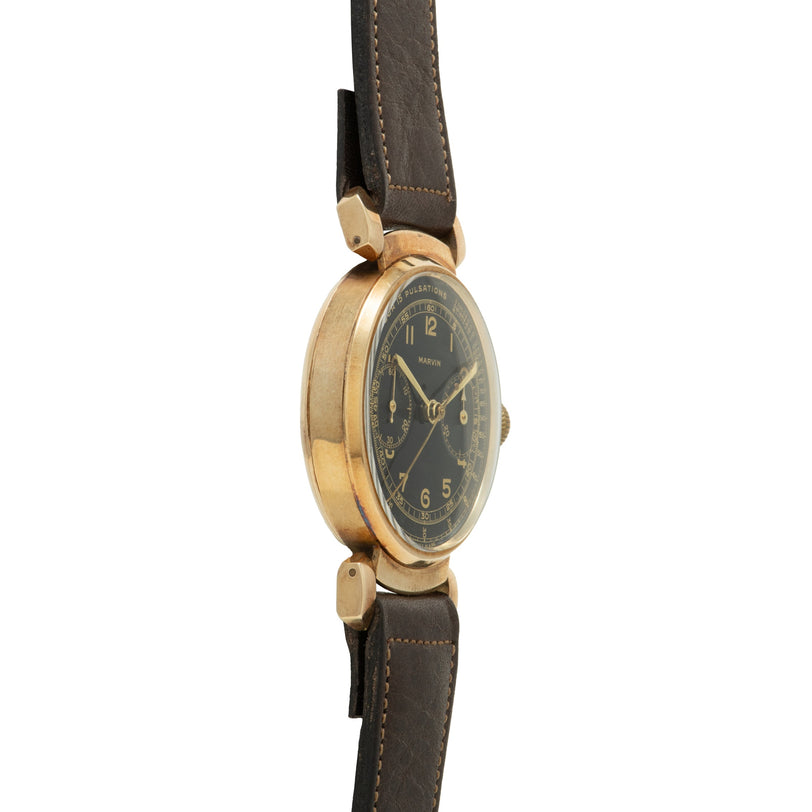 Gold-tone chronograph watch with black dial, Arabic numerals, and dark leather strap with hinged lugs.