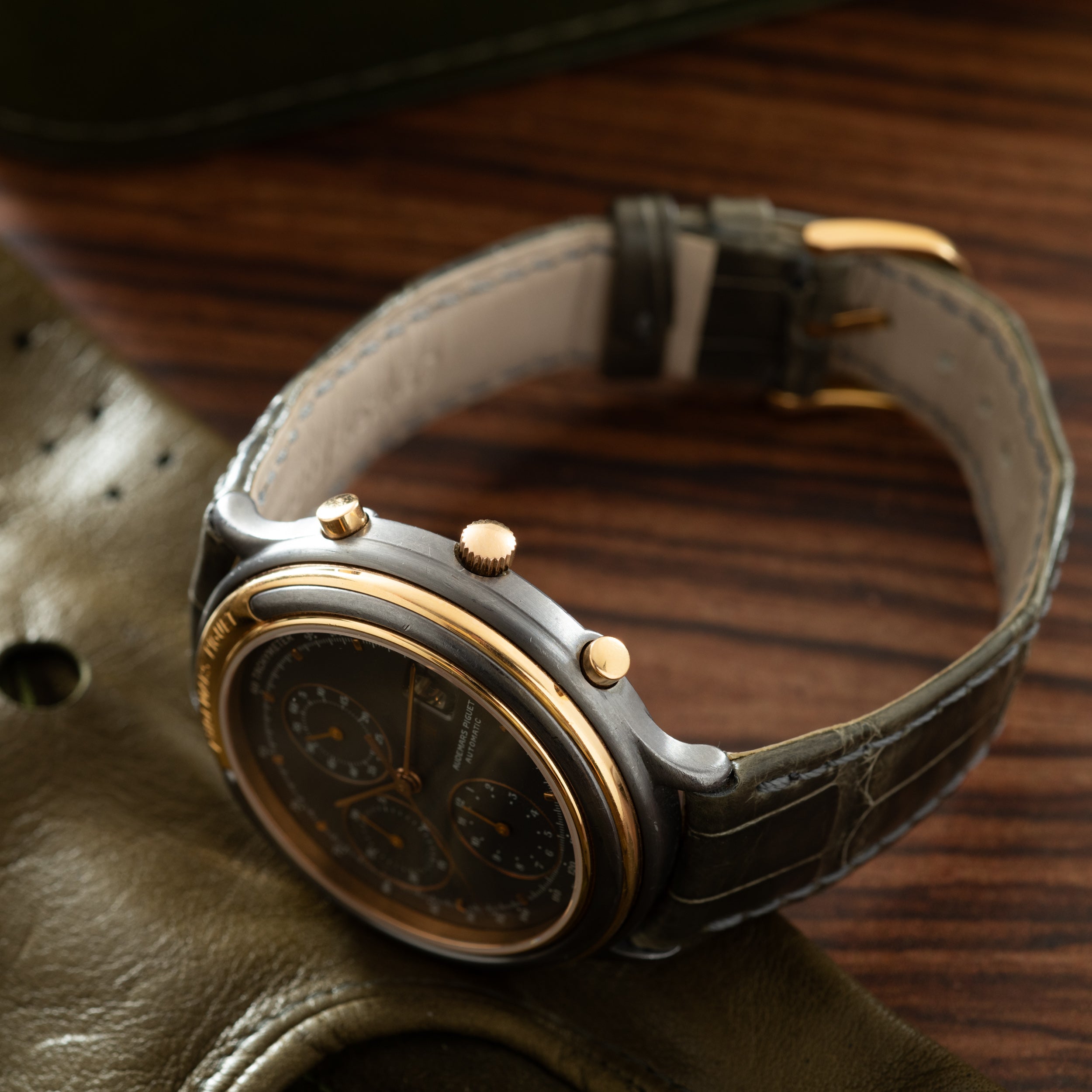 Steel and rose gold chronograph watch with black dial and dark leather strap on wooden surface.