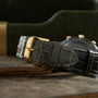 Black crocodile leather strap with gold-tone buckle on chronograph watch with dark dial and gold pushers.