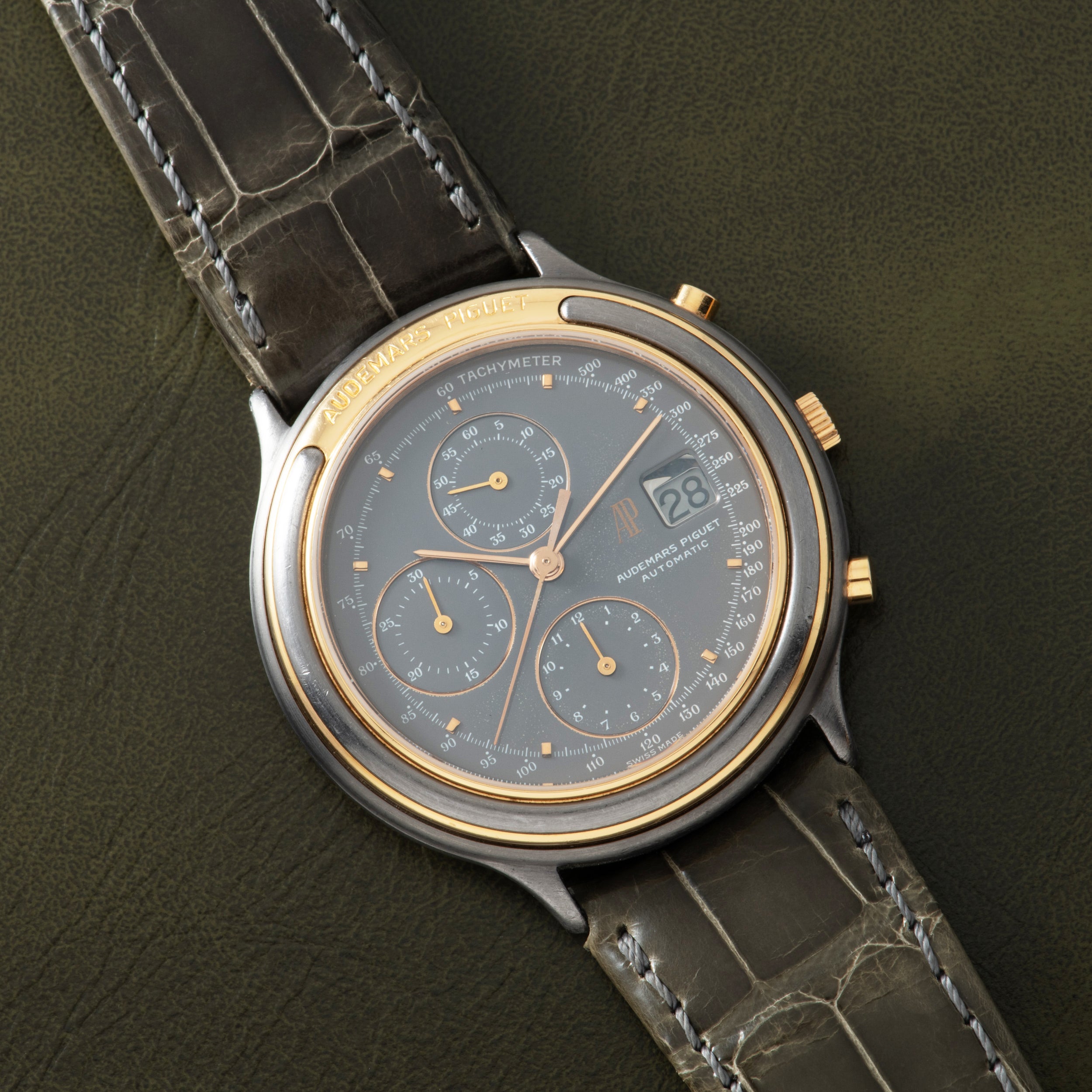 Stainless steel and gold-tone chronograph watch with gray dial, three subdials, and dark leather crocodile-embossed strap.