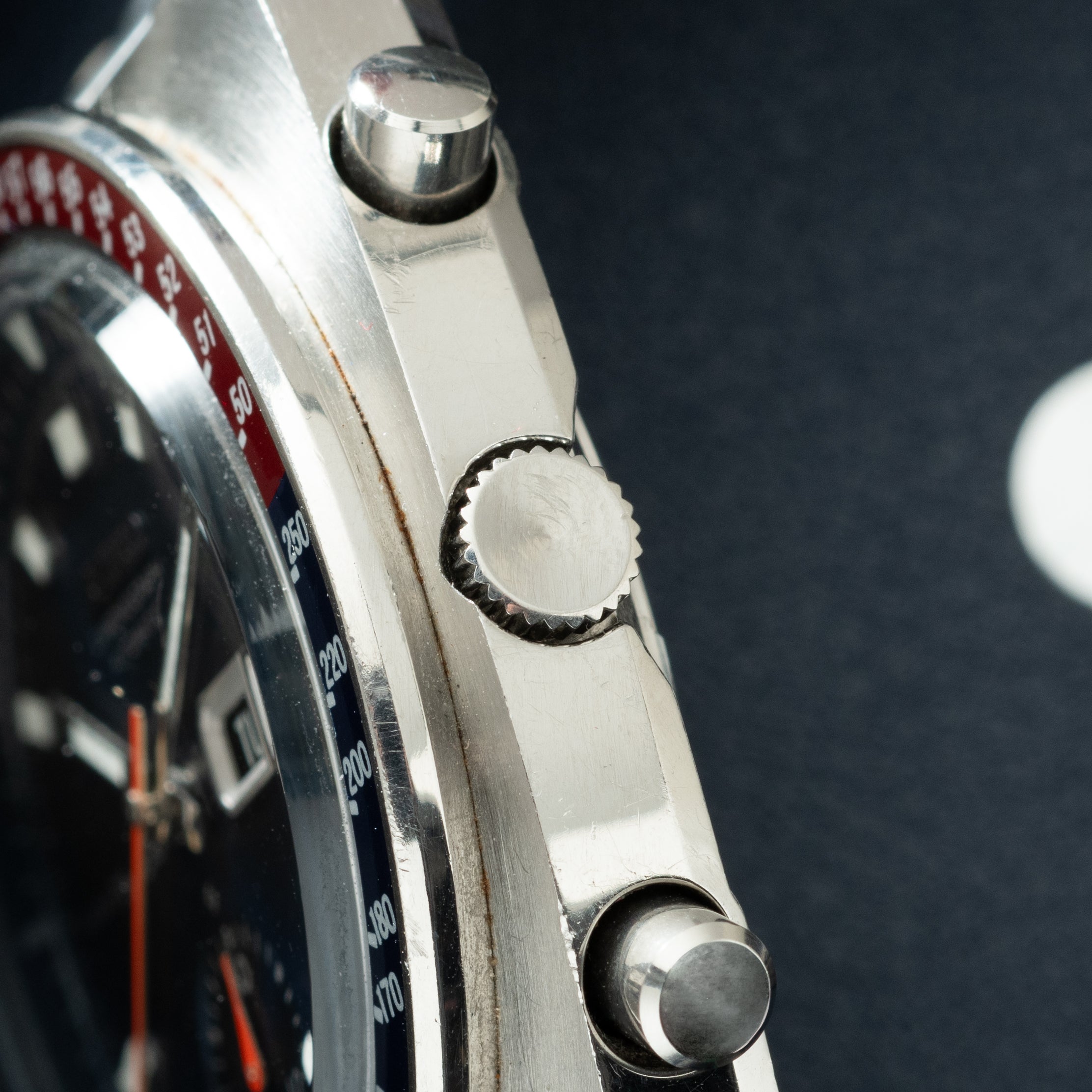 Silver chronograph watch with red and black bezel, showing side profile of case, crown, and pushers against dark background.