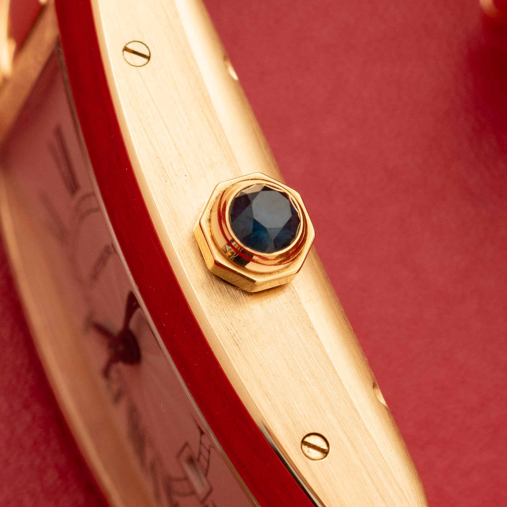 Gold watch bracelet with dark blue faceted stone crown and screw details on brushed gold band.