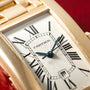 Gold Cartier Tank Américaine watch with white dial, Roman numerals, and blue hands on red background.